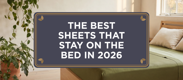Blog header crest reading The Best Sheets That Stay On the Bed in 2026 over a warm modern-minimal plant-forward bedroom with an oak bed dressed in Olive Green organic bamboo viscose One Sheet in soft afternoon light